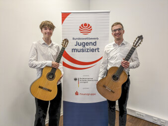zwei Jugendliche mit Gitarren neben einem Aufsteller mit Schriftzug "Jugend musiziert"