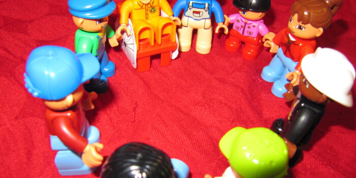Unterschiedlich bemahlte kleine Spielfiguren, darunter eine Figur in einem Rollstuhl, stehen sich an den Händen haltend in einem Kreis auf einer roten Decke