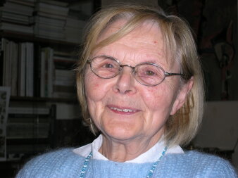 Eine ältere, lächelnde Dame mit blonden Haaren, Brille und blauem Pullover.