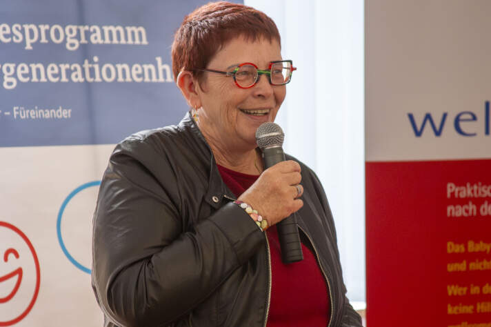 Eine Frau mit kurzen, roten Haaren und bunter Brille hält ein Mikrofon in der Hand und spricht hinein. 