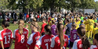 Viele Kinder in Fußballtrikots stehen auf einer Wiese. Vor ihnen spricht eine Frau in ein Mikrofon. 