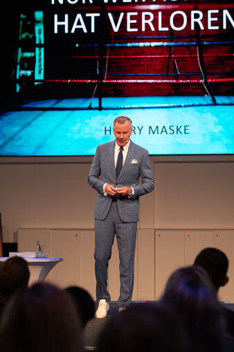 Ein Mann steht auf einer Bühne und hält einen Vortrag. Im Hintergrund läuft eine Präsentation.
