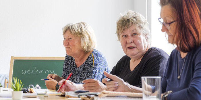 Drei ältere Frauen sitzen an einem Tisch. Im Hintergrund steht eine Tafel. Darauf steht das Wort „welcome“.