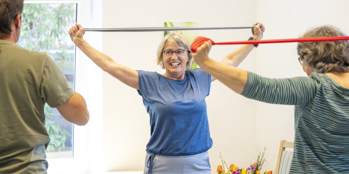 EIne Frau zeigt zwei anderen Personen eine Sportübung mit einem Band